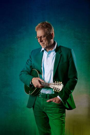 Eric Chasalow looks down contemplatively as he holds a stringed instrument. He stands in front of a multicolored background.