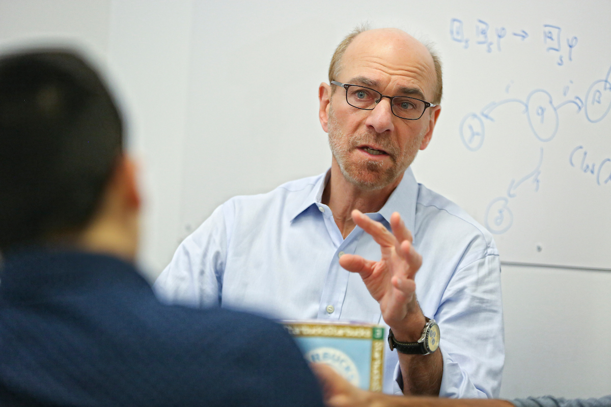 computer science professor James Pustejovsky speaks in front of a white board with blue writing on it. The perspective is over someone's shoulder.