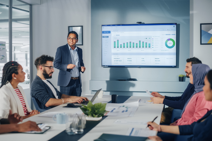 man stands in front of a leadership table, pointing at graphs and statistics