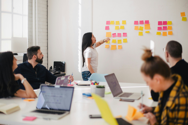 Woman points at sticky notes on a whiteboard while giving a presentation.