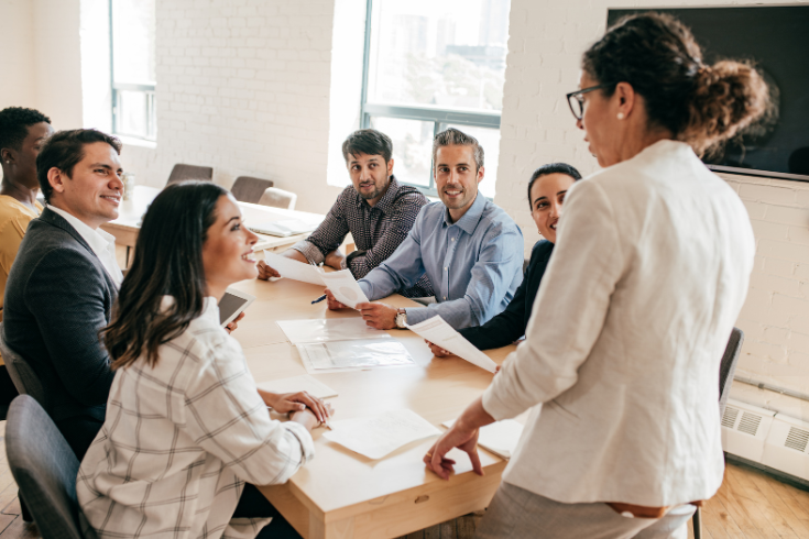 Person standing at the head of a conference table, talking to coworkers