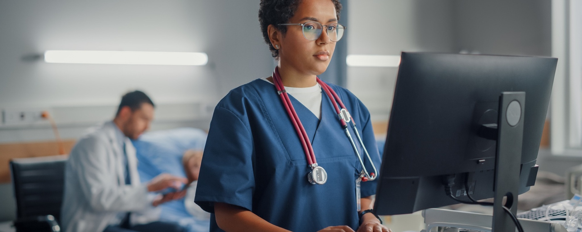 medical professional looking at computer with doctor and patient blurred in background. 