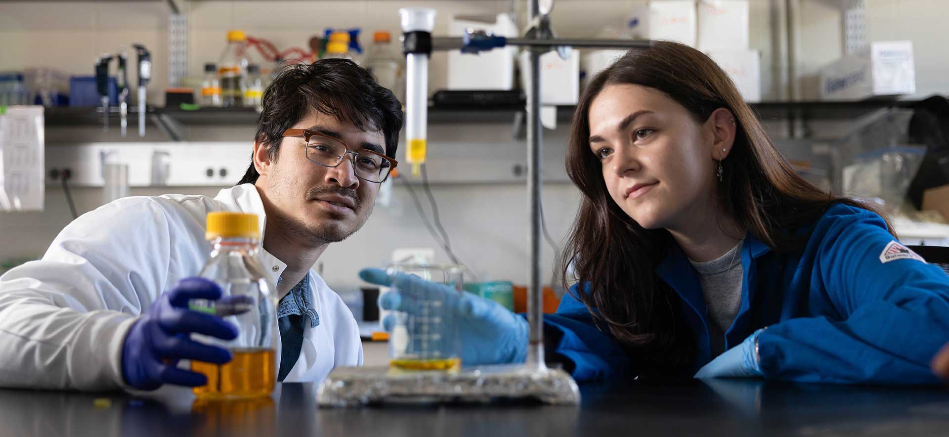 A researcher wearing blue gloves and safety goggles pours a clear liquid into a test tube