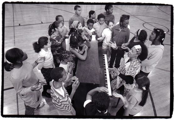 Children surround Jane Sapp while she is playing piano