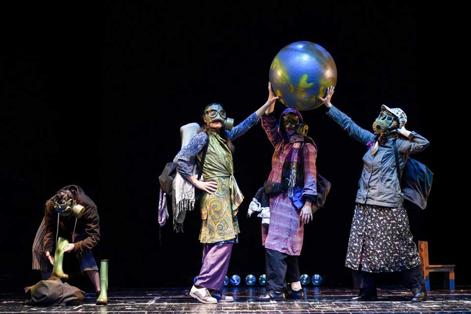 Actors in gas masks stand on a stage holding up a ball that looks like Earth