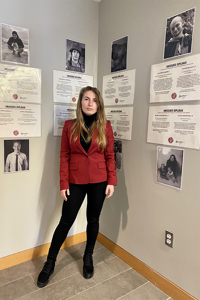 A woman stands in a corner of a room, wearing a red blazer over a black turtleneck and black pants. Behind her on the walls are several printed pages titled "UNISSUED DIPLOMA," accompanied by black-and-white photographs of young individuals, suggesting a commemorative or memorial display. The floor is tiled, and there is a visible electrical outlet on the right wall. The atmosphere is solemn and reflective.