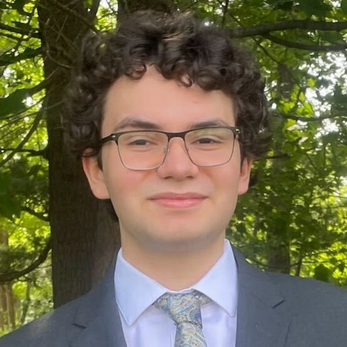 a young man with glasses in a tuxedo smiling with trees in the background. this is a headshot