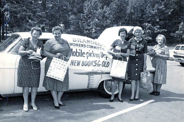 Black and white photo of five women holding bags and books, next to a car used to collect books
