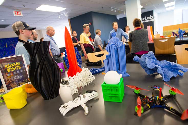 Colorful objects in the foreground, products of 3-D printing. People observe on a tour in the background.