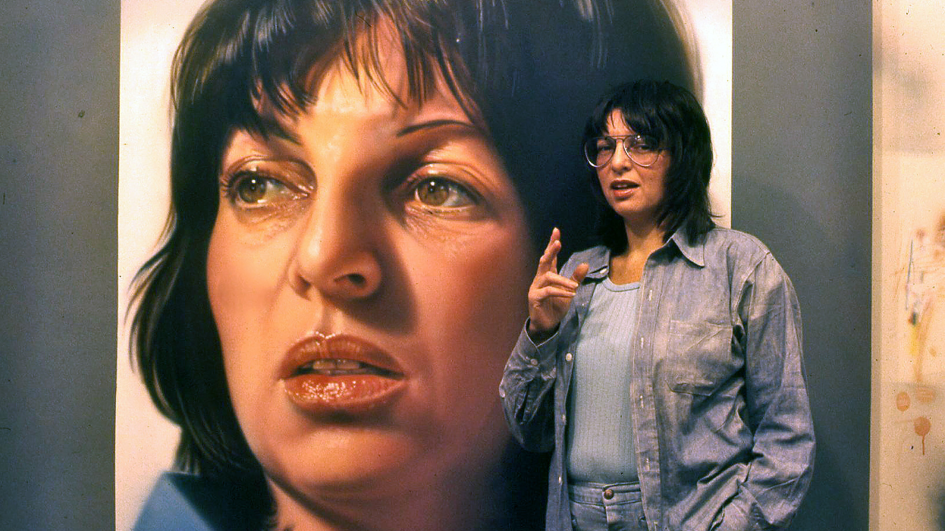 Film still from Queen of Hearts showing artist Audrey Flack standing in front of a self-portrait
