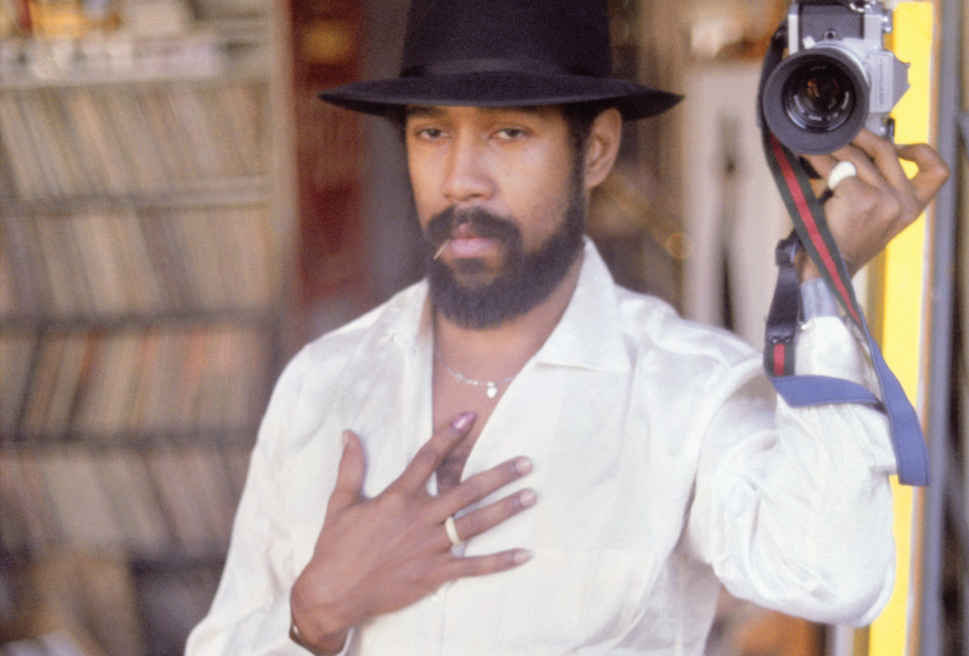 Self-portrait of Barkley L. Hendricks in a white shirt, wearing a black hat and holding a camera