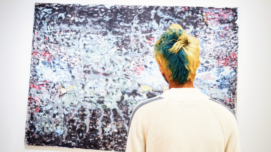 Young person with yellow and greenish-blue hair looking at an abstract painting by Mark Bradford.