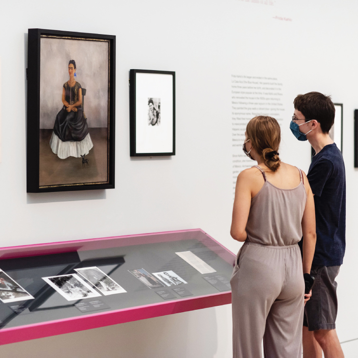 A young white woman and white man look at the Frida Kahlo "Itzcuintli Dog with Me" which features a seated self-portrait of Frida Kahlo with a small dog.