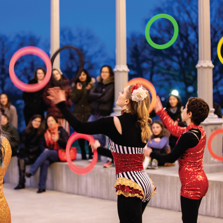 Four circus performers throw rings into the air in front of a crowd of spectators.