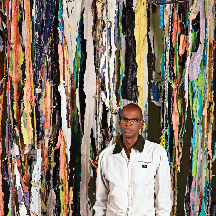 A Black man wearing black glasses and a white sweater standing in front of strips of painted canvas.