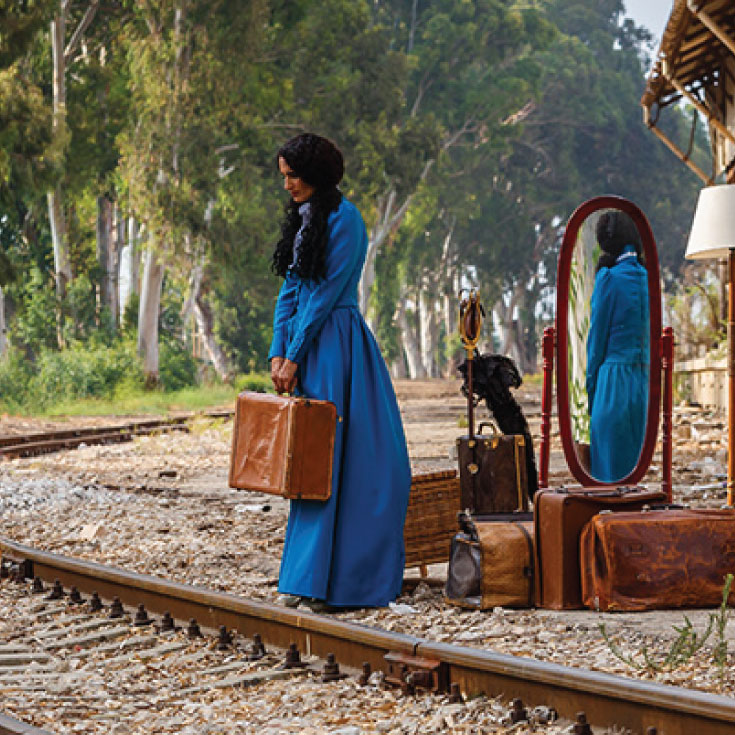 A woman in a blue dress holding a suitcase standing next to a railroad track.