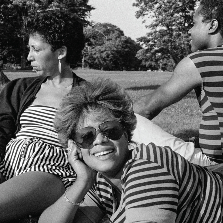Two Black women and one Black man lounge on the grass in a black and white photograph by Barkley L. Hendricks.