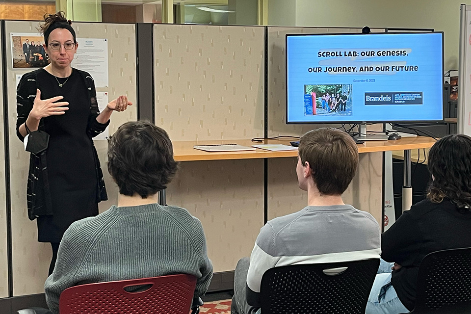 Professor Hassenfeld stands addressing a group of students seated around a large wooden table with many black chairs around it and a patterned rug underneath. In the background, there is a whiteboard, a screen with students on Zoom, a lamp, and a plant. All people are wearing masks. The table is covered in laptops and papers.