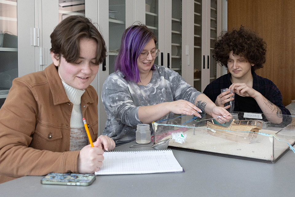 Three students inspect a miniature artifact excavated from a handmade model of a shipwreck