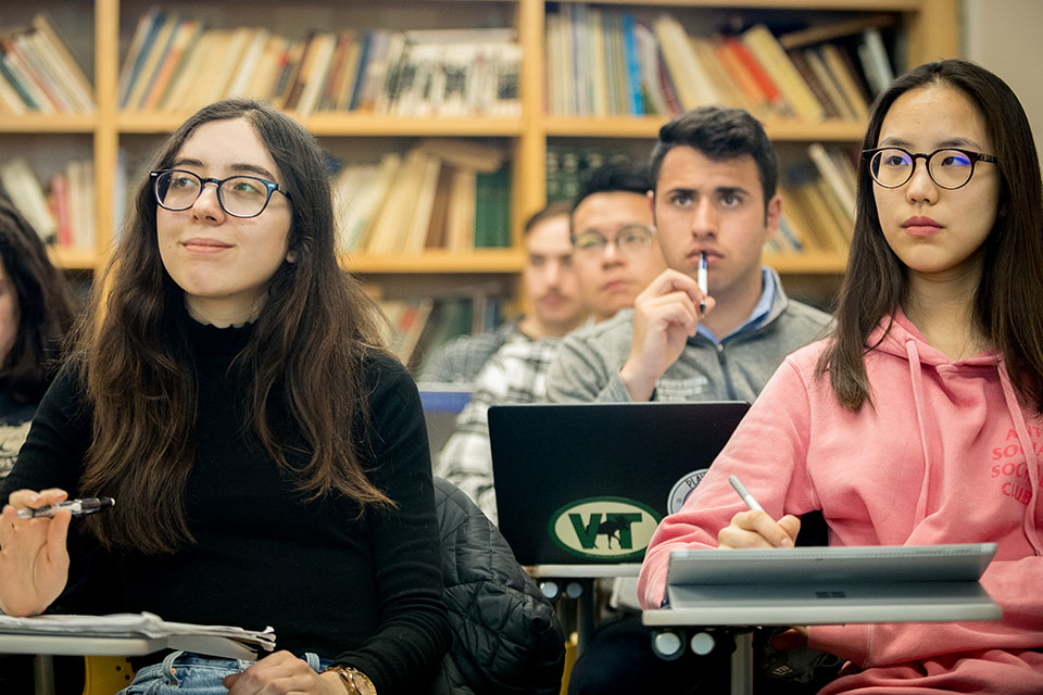 Students sitting in classroom desks with a bookshelf behind them.