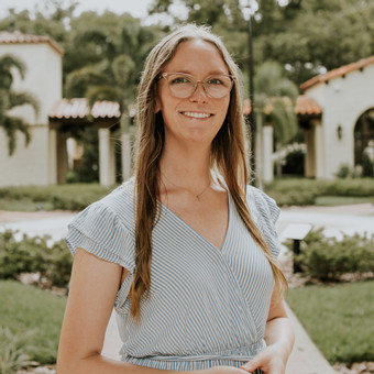 picture of woman with long straight brown hair, large glasses and a striped dress smilng in front of a building with red tiled southwestern roof