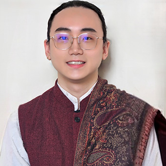 smiling young adult with thin gold glasses and black hair pulled back, wearing dark red patterned cloth over white shirt