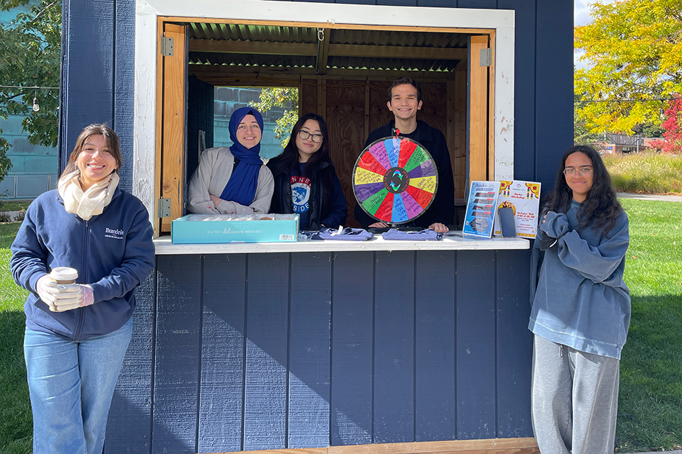 5 students smiling at the camera from inside and out of a Brandeis blue booth, with a spinner and small things in front of them