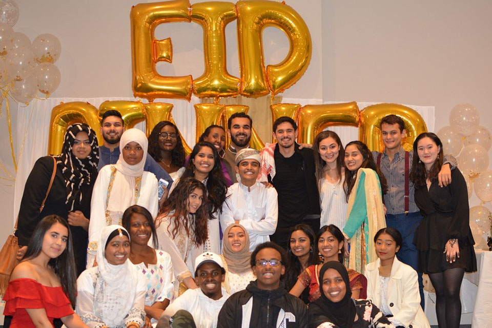 male and female smiling students, dressed festively and some wearing headcoverings, stand together under a balloon sign that reads "Eid Dinner"