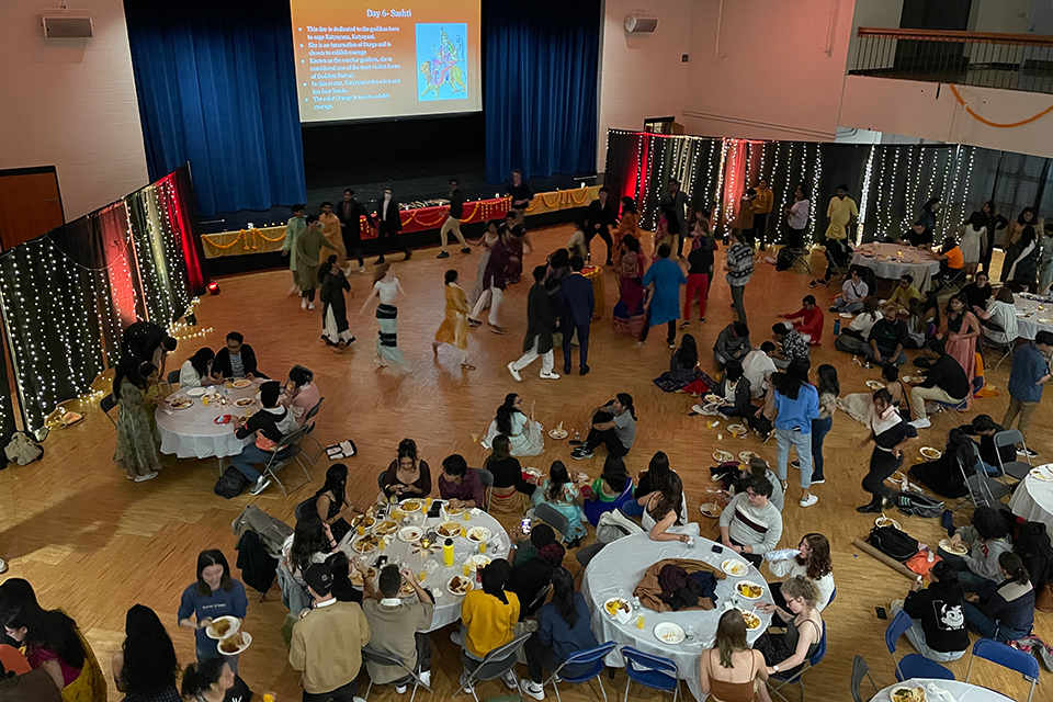 A large room fille with costumed people, some circling in dance, and a large screen in the background pulled down from the ceiling with an illegible slide. The scene is very colorful.