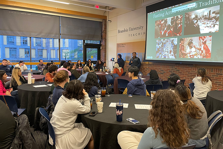 students sitting around tables with black-haired man speaking next to a large screen with a picture of people and words about fasting across traditions