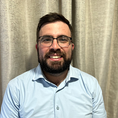 A headshot of a broadly smiling man with dark hair, glasses, and short beard