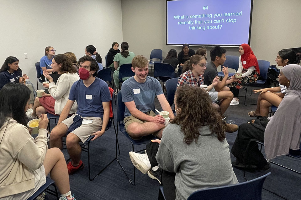 a large group of smiling students stand facing the camera in front of a screen that says Center for Spiritual Life. A dog is sitting and facing the students wearing a tshirt similar to ones many students are holding up. Students wear nametags and a couple women wear hijabs