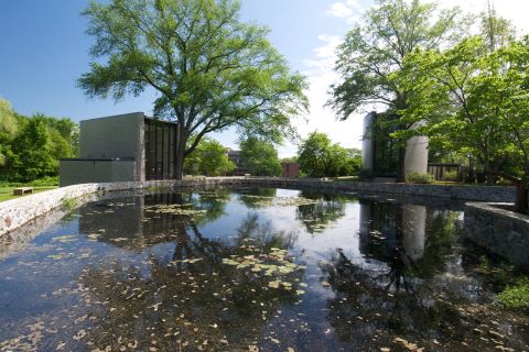 photo of a manmade pond with a brick wall around it in the foreground and two brick chapels in the background with trees next to and behind the chapels, with young green leaves