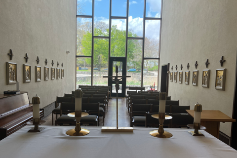 photo of altar in foreground with crucifix and chairs behind, with tall window in the background with cars visible parked in the distance