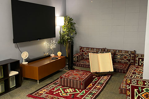 view from one room with Muslim prayer rugs on the floor and Muslim script on the wall; through open wooden doors is a room with a couch and a door going into an inner office