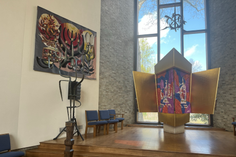 Colorful Jewish Ark holding a Torah with colorful curtain in foreground, an elaborate candlestick and a wool wall weaving on the left and tall windows in the back