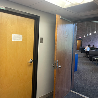 There is a closed door on the left of this image, and an open door on the right, through which one can see tables with seats, blue carpeting and lamps hanging overhead