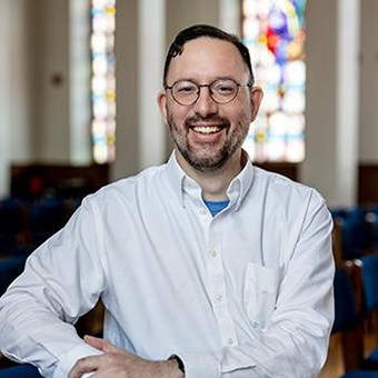 a picture of a young white man with glasses and slight mustache and beard smiling broadly and sitting on and near blue chairs with stained glass windows behind