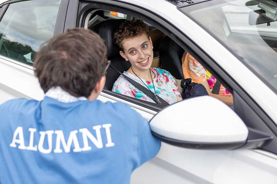 Person in a car speak with someone in a blue shirt that reads, Alumni.