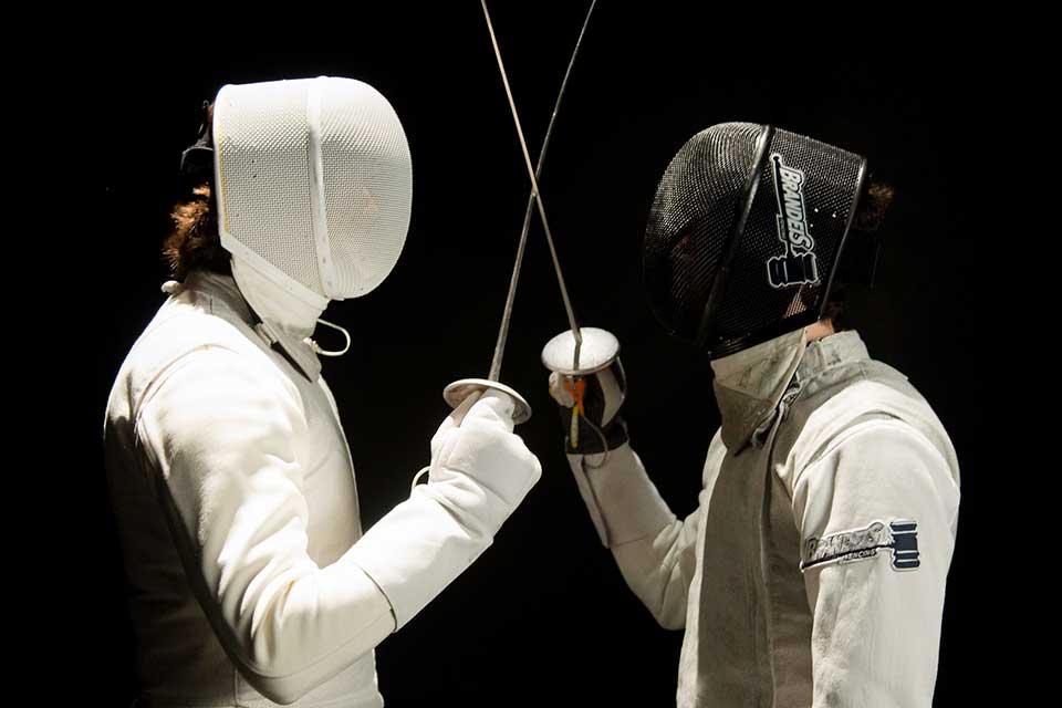 Two fencers in helmets look at each other closely with swords crossed
