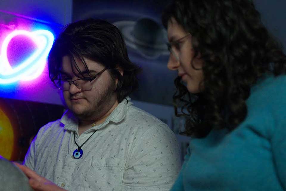 Two people looking at something with a planet-shaped neon sign behind them