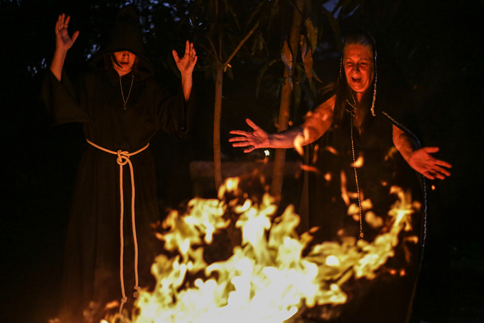 Two people stand around fire.