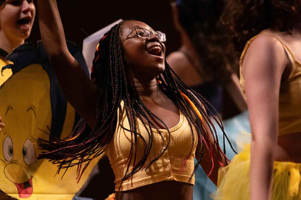 Person dressed in yellow performs on stage, a person wearing a Flounder fish costume is seen in the background