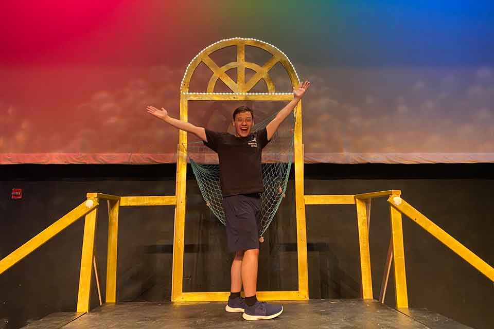 Person stands on stage with arms outstretched in front of a colorful backdrop