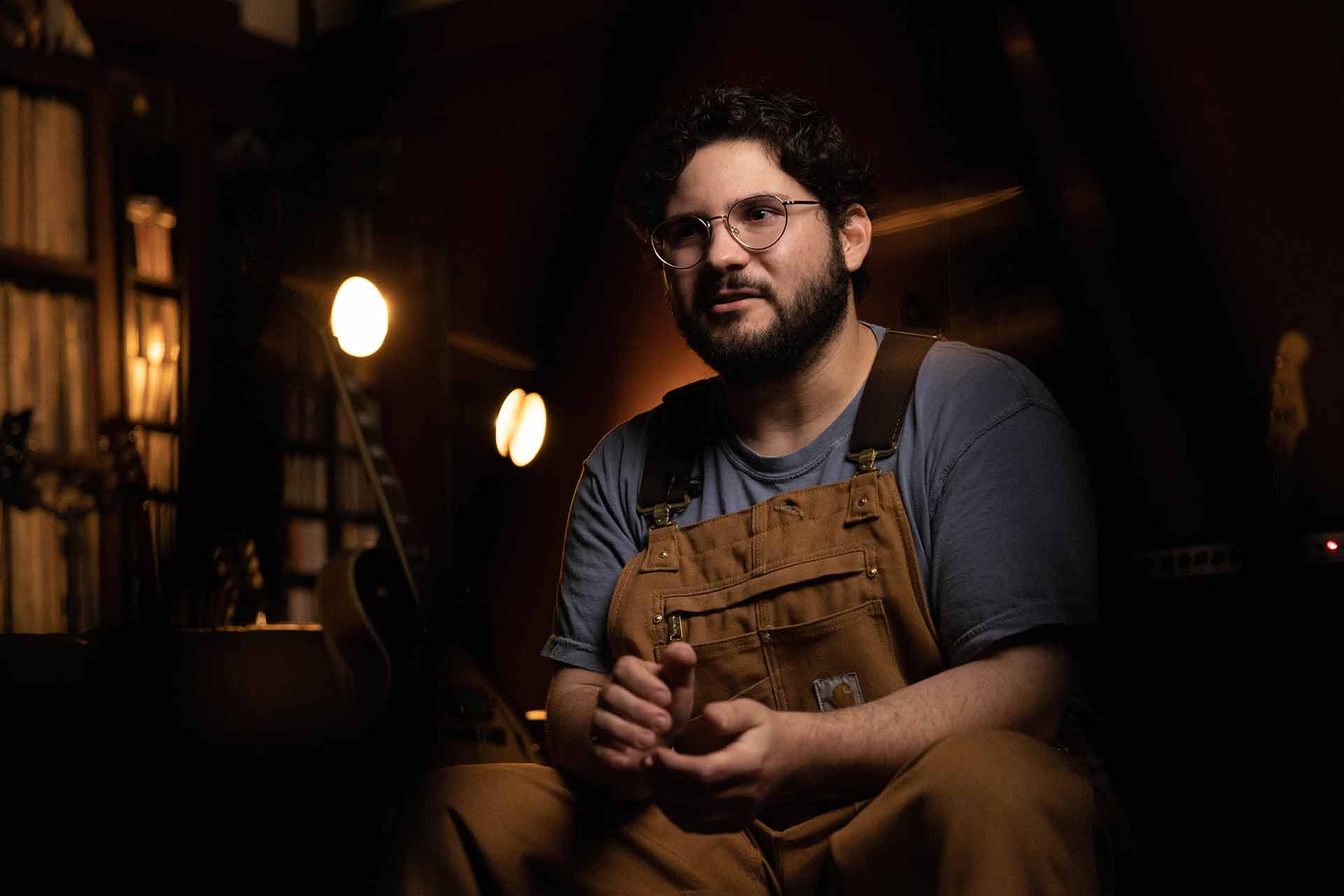 Sam is wearing brown overalls and is sitting. Two lights shine down on him in the background.
