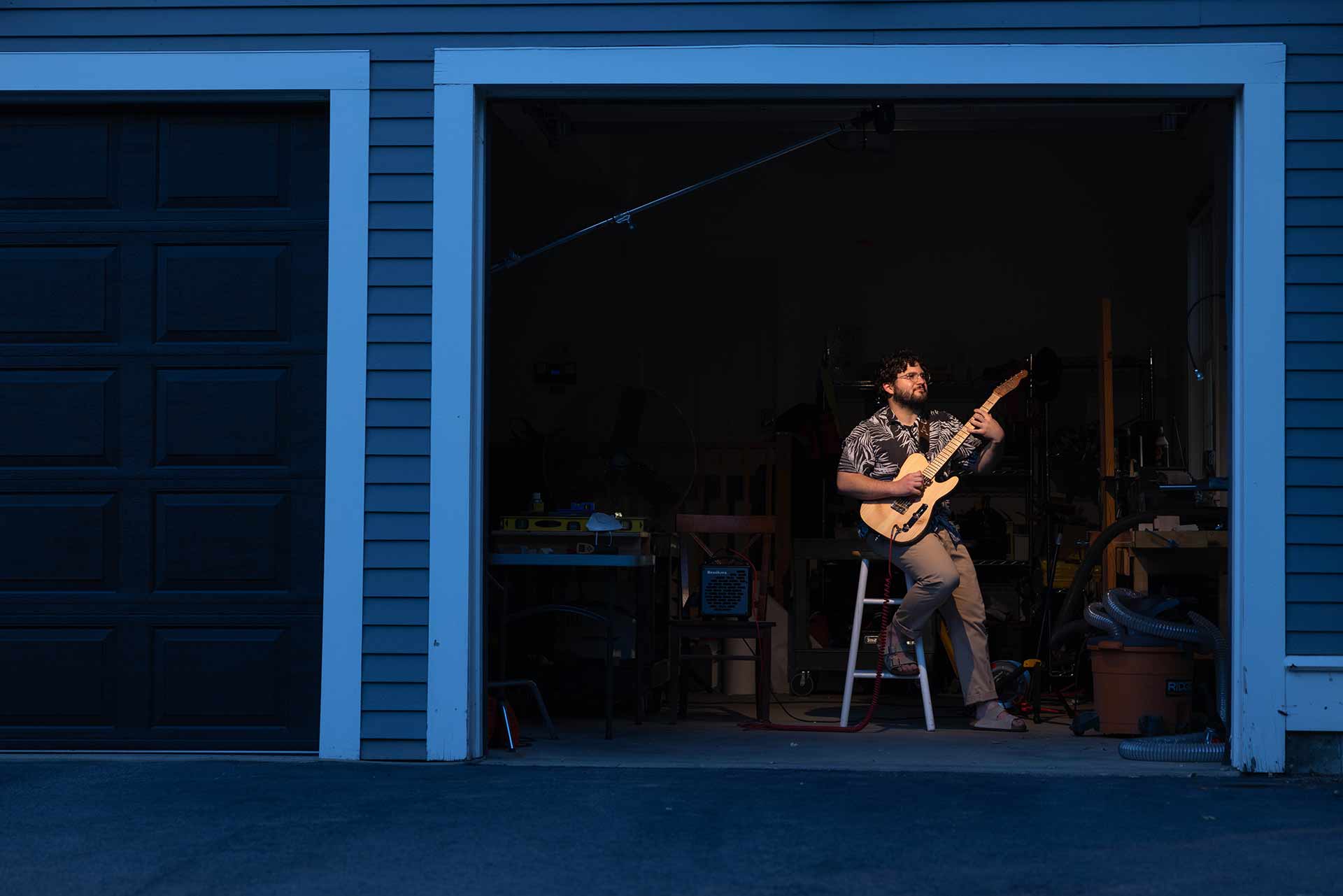 Sam sits on a stool, framed by the door of his garage. Light shines on him as he plays a guitar.