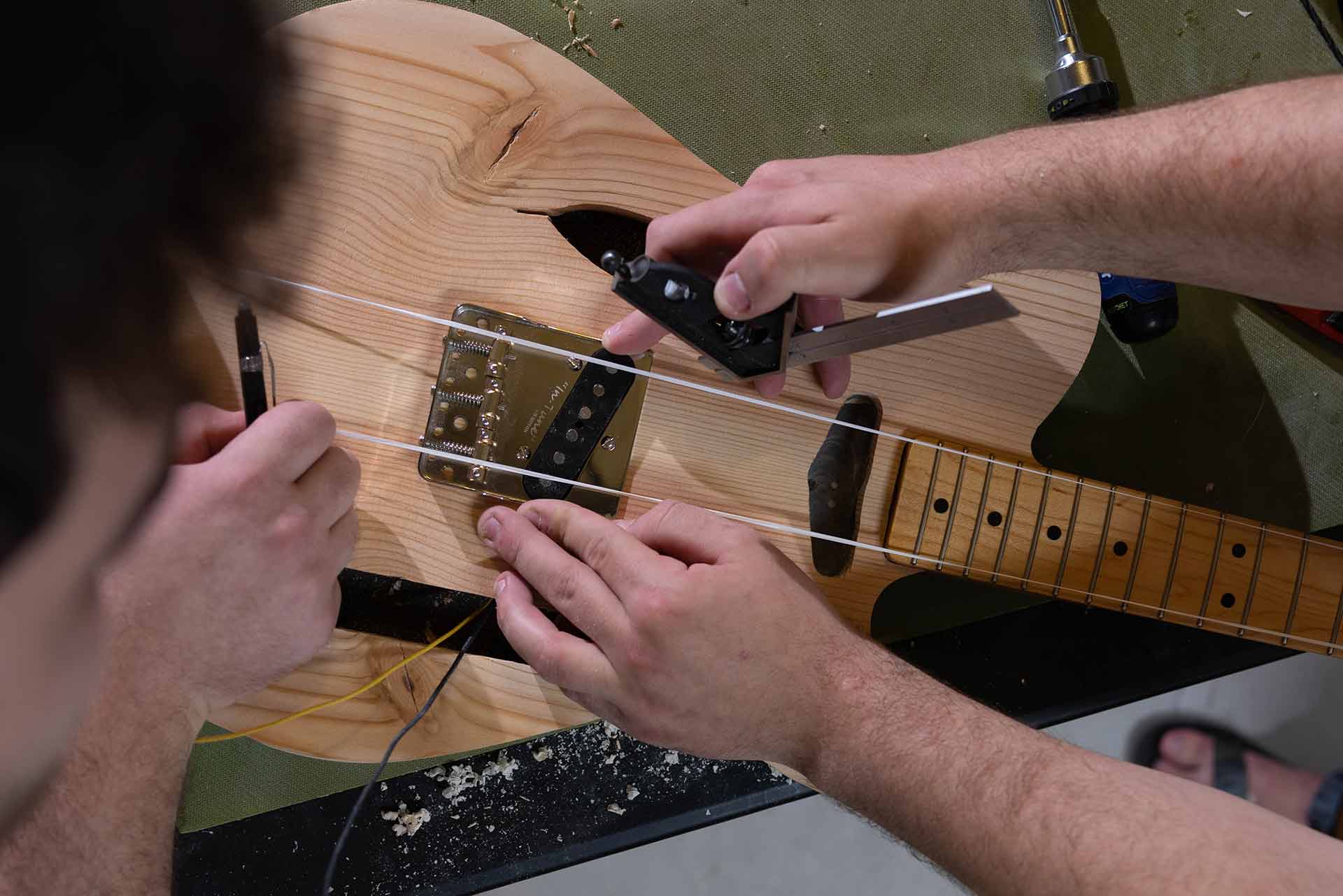 Two people take measurements while building a guitar. One holds a pen and one holds a square (tool).