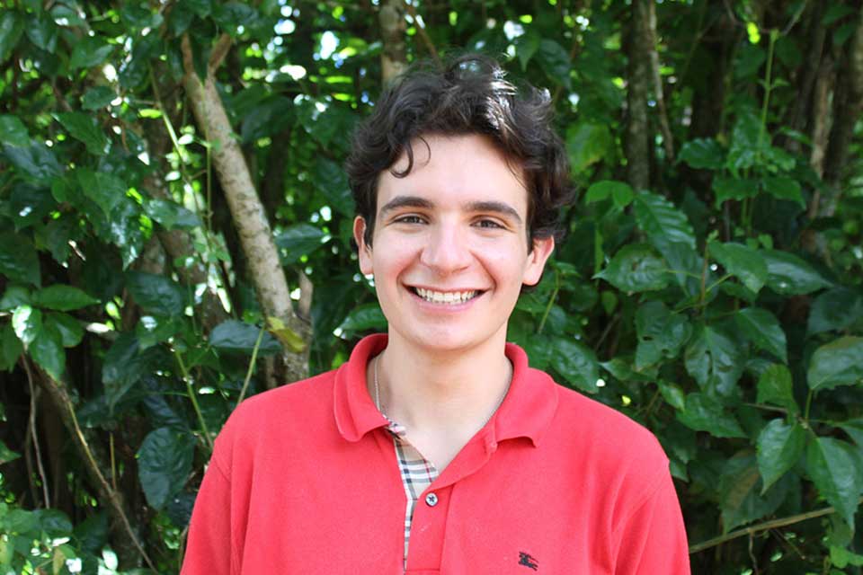 Daniel Block, a young man with dark hair that hangs in loose curls,  standing in a collared red shortsleeve shirt in front of a leafy bush.