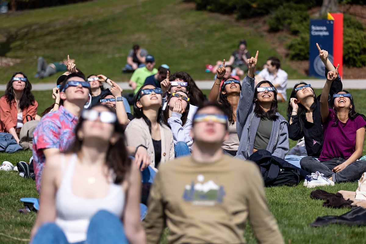 Students seated on the grass look up toward the sky wearing eclipse glasses