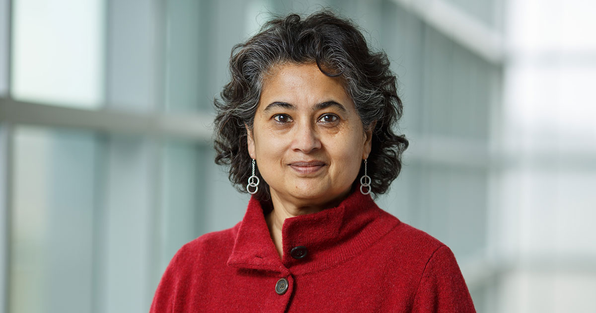 Head shot of piali sengupta, wearing a red sweater and dangling silver earrings, staring directly into the camera.